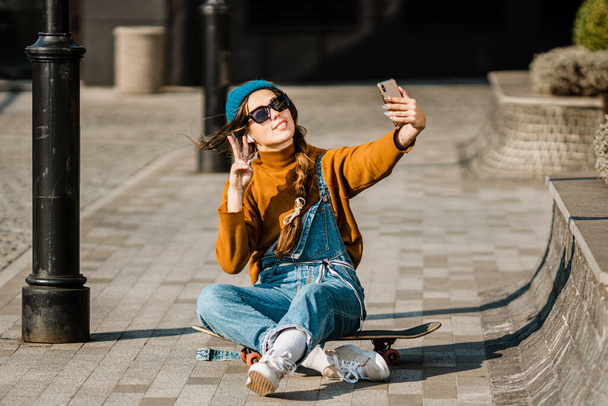 Cute urban girl outdoors with skateboard using smart phone. Happy young woman with skateboard, headphones and cell phone. Hipster female sit on skate board and using mobile phone. - Photo, Image