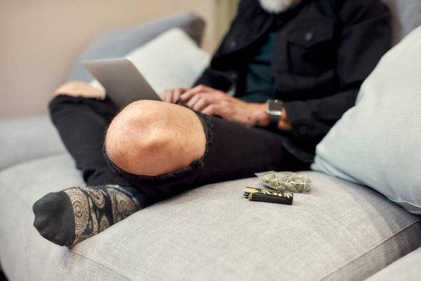 For higher productivity. Close up of buds in plastic bag and lighter lying on the couch. Man using laptop in the background. Cannabis and weed legalization concept - Photo, image