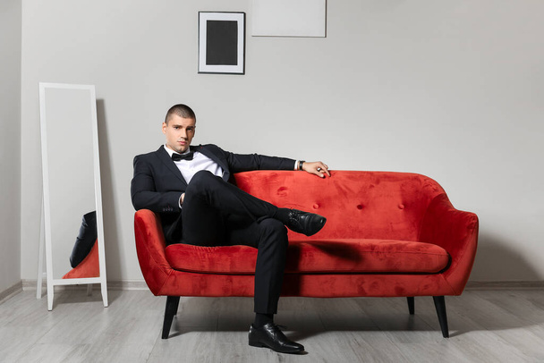 Όμορφος νεαρός άνδρας με κομψό κοστούμι κάθεται στον καναπέ σε εσωτερικούς χώρους - Φωτογραφία, εικόνα