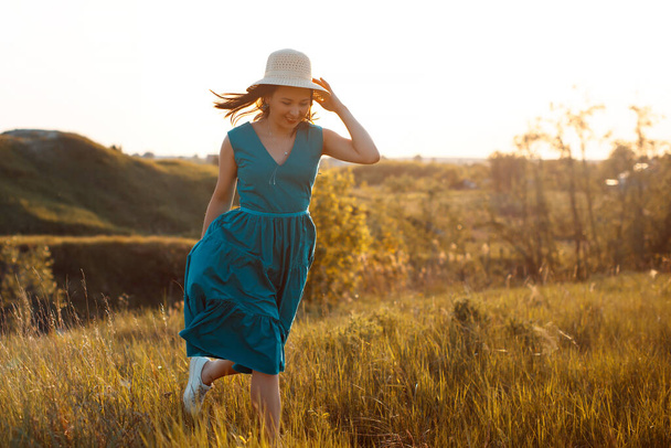 woman in a blue dress and panama runs on a summer meadow at sunset. The concept of outdoor recreation, youth, summer, freedom, health - Foto, Bild