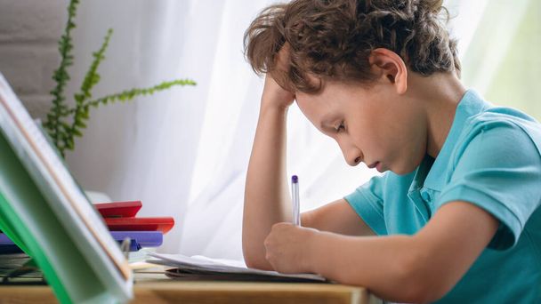 sad schoolboy puts his hand under his head and looks at a notebook while sitting at a Desk and doing homework. at home. Back to school. The child is tired of studying. End of the school year. - Foto, afbeelding