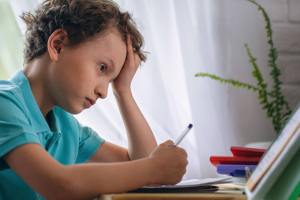 sad schoolboy puts his hand under his head and looks at book, sits at Desk and reads homework. Back to school. child is tired of studying. End of school year. child does not understand the task topic. - Foto, Imagem