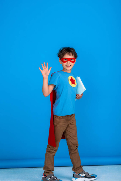 cheerful child dressed in superhero costume holds a book and waves. little boy laughs and enjoys learning at school. Posing on pink background in Studio. Forward to knowledge, back to school. - Photo, image