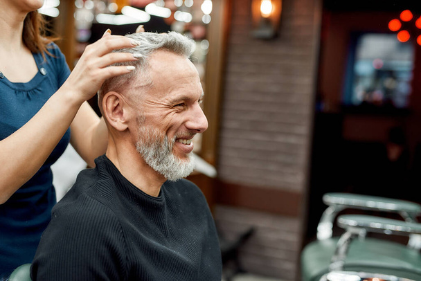 I like it. Happy mature bearded man smiling while female barber making haircut for him, he is sitting in barbershop - Fotó, kép
