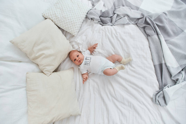 Söpö 4 kuukauden ikäinen vauva valkoisessa kehonpuvussa ja sukissa, makaa selällään sängyllä ja katsoo kehykseen. Sänky kevyillä lakanoilla, huopilla ja tyynyillä. Onnellinen pikkumies. näkymä ylhäältä
 - Valokuva, kuva