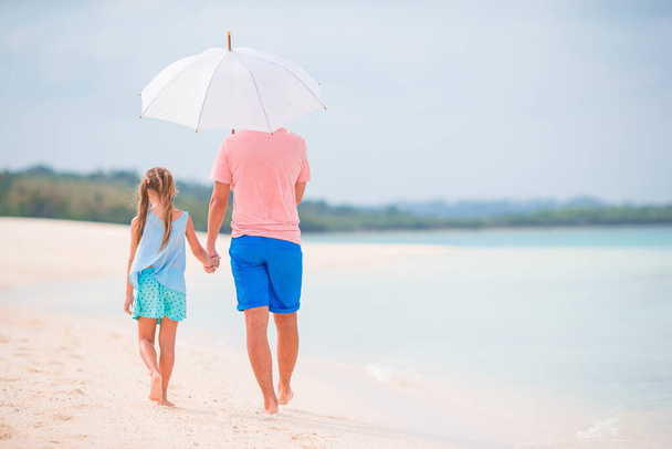 Young father and his little daughter walking under umbrella on white sand beach - Foto, Imagem