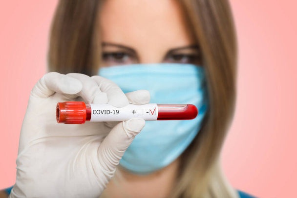 Nurse doctor in medical mask holding test tube with positive blood test result for Coronavirus on blured background. Test tube with blood and inscription on tag covid-19. Laboratory studies - Photo, Image
