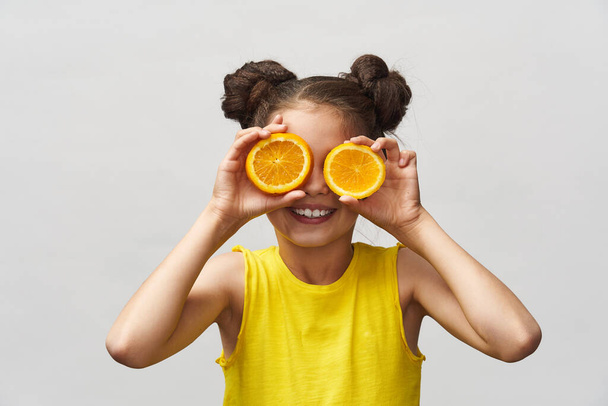 Cute charming little girl in a yellow t-shirt, laughing merrily and holding a half orange on a light background. The child is Caucasian. Concept of summer and vitamins. - Foto, Imagen