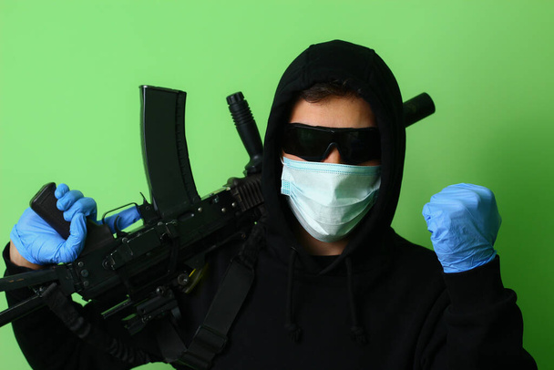 Jovem gangster masculino ou criminoso em uma máscara cirúrgica médica e óculos de sol vestidos com roupas pretas com um capuz detém uma arma de assalto automático
 - Foto, Imagem
