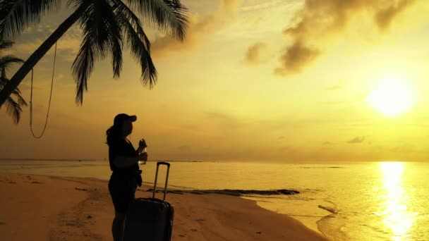Nainen seisoo trooppisella rannalla matkalaukku auringonlaskun aikaan - Materiaali, video