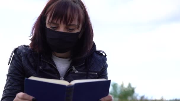 Close up of young woman in black medical mask and casual clothes sitting on hill and reading book on cloudy day. Adult female resting alone in countryside. - Séquence, vidéo