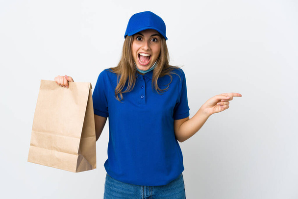 Food delivery woman and protecting from the coronavirus with a mask over isolated white background surprised and pointing finger to the side - 写真・画像