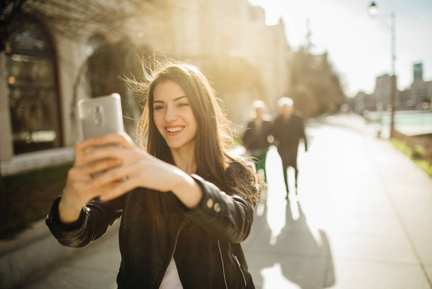 Young attractive woman making selfie outdoors.Woman taking smartphone self portrait with front camera.Sharing pictures on social media.Smiling girl walking on city streets talking on video call - Foto, afbeelding