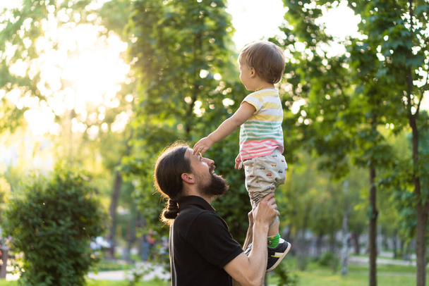 Fatherhood, parenthood, childhood, caring, summer and leisure concept - young dad with beard and long hair in black t-shirt holds in his arms little son in the backlight of the sunset in the park. - Φωτογραφία, εικόνα