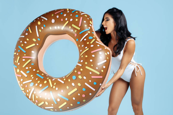 Excited fit young woman dressed in swimsuit posing happy with donut inflatable circle over blue background. - Foto, immagini