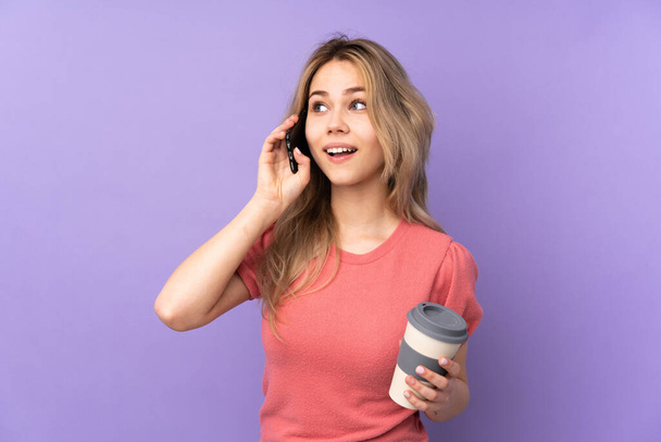 Teenager russische Mädchen isoliert auf lila Hintergrund hält Kaffee zum Mitnehmen und ein Handy - Foto, Bild