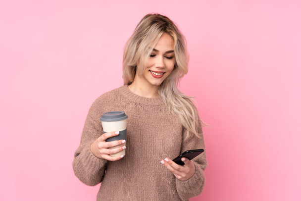 Tini szőke lány visel pulóver felett elszigetelt rózsaszín háttér kezében kávét, hogy vigye el, és egy mobil - Fotó, kép