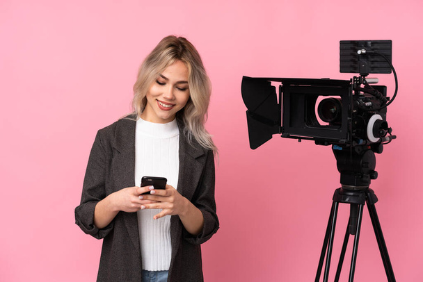 Женщина-репортер держит микрофон и сообщает новости на изолированном розовом фоне, отправляя сообщение с мобильного телефона - Фото, изображение