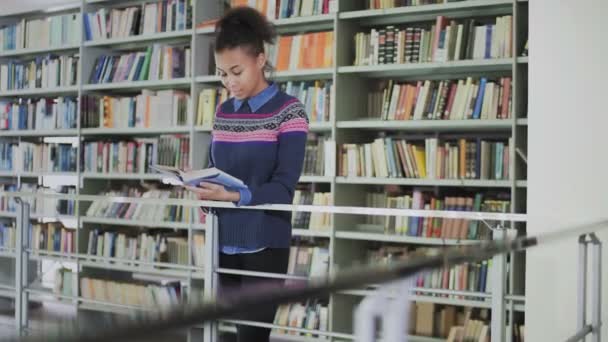 Portret van de jonge krullende Afro-Amerikaanse vrouw die het oude boek leest terwijl ze bij de boekenplanken in de bibliotheek staat. - Video