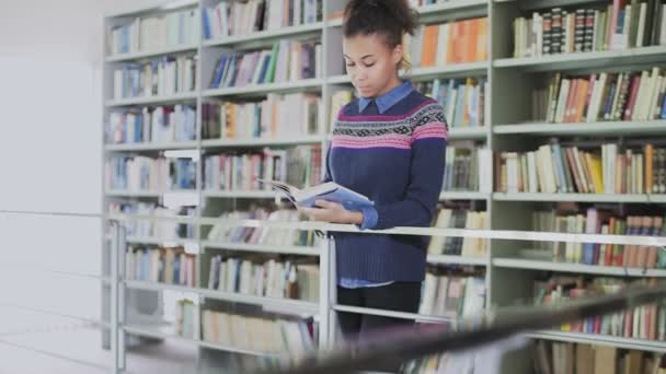 Dolly Bewegung der jungen lockigen Afroamerikanerin beim Lesen des alten Buches, während sie in der Nähe der Bücherregale in der Bibliothek steht. - Filmmaterial, Video