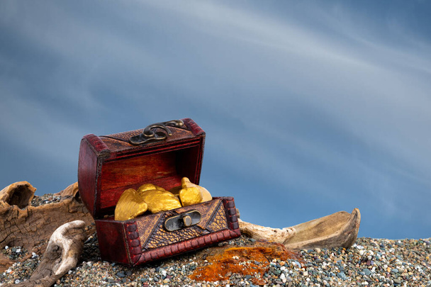 A small treasure chest filled with gold nuggets. Chest is on a sandy beach with driftwood. Background is blue sky with clouds. - Photo, Image