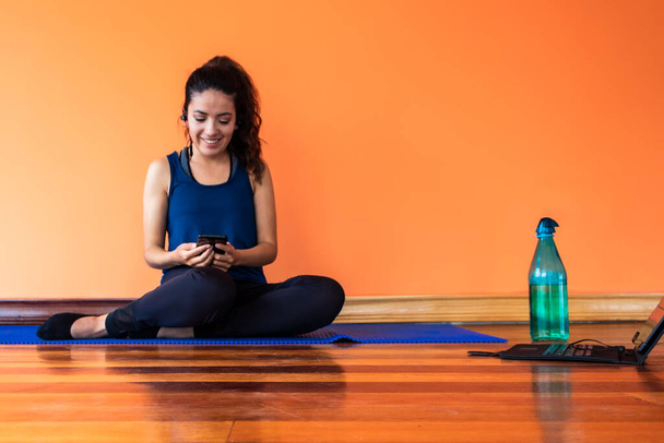 Jonge vrouw zittend op een blauwe mat kijkend naar haar mobiele telefoon, met behulp van draadloze koptelefoon en glimlachend - Foto, afbeelding
