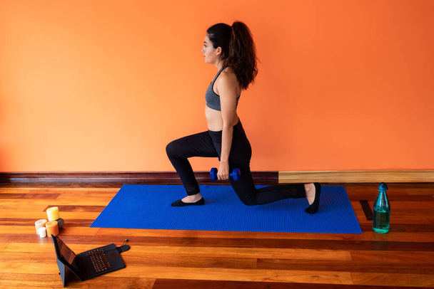 jonge vrouw doet een squat met behulp van blauwe halters, staande op een mat - Foto, afbeelding