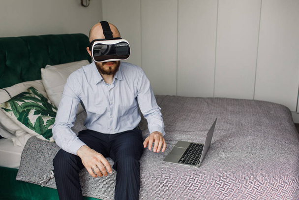 young man with a beard in virtual reality glasses sits on the edge of the bed - Valokuva, kuva