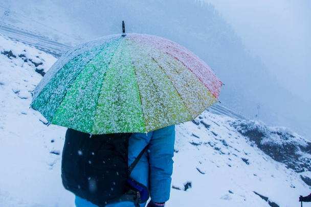Πρόσωπο στέκεται με πολύχρωμη ομπρέλα ουράνιο τόξο, πτώση χιονιού, Λευκό τοπίο και παχύ χιόνι το χειμώνα αναζητούν παγωμένο και κρύο, διακοπές, ο τουρισμός ξεκίνησε κατά τη διάρκεια covid-19 - Φωτογραφία, εικόνα