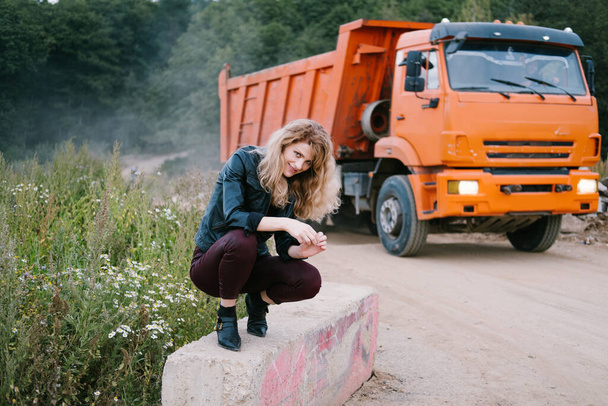 Woman wearing leather jacket seating near field road with big truck. Industrial Zone - 写真・画像