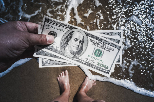 Девушка с деньгами в 300 долларов на фоне морских океанских волн с белой пеной и песчаным мокрым пляжем с ногами в песке крупным планом. Концепция финансирования отпуска деньги путешествия доллары отпуск - Фото, изображение