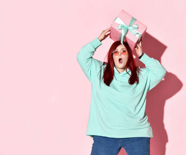 Happy surprised overweight woman in blue jeans, hoodie and sunglasses holding gift box on her head - Fotoğraf, Görsel