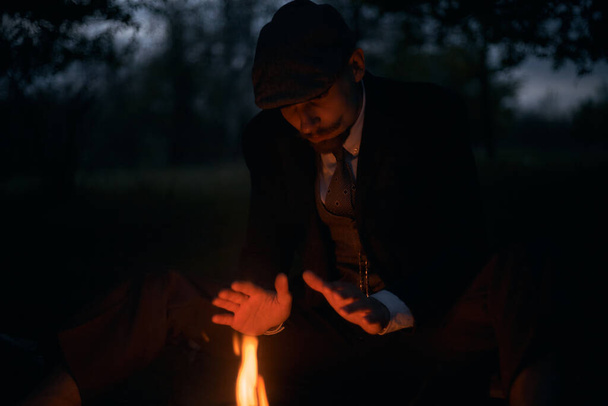 Ritratto di un ragazzo seduto accanto al fuoco di notte nella natura. Foto d'epoca cinematografica. Ritratto retrò anni '20 di un uomo inglese con un cappuccio piatto
. - Foto, immagini