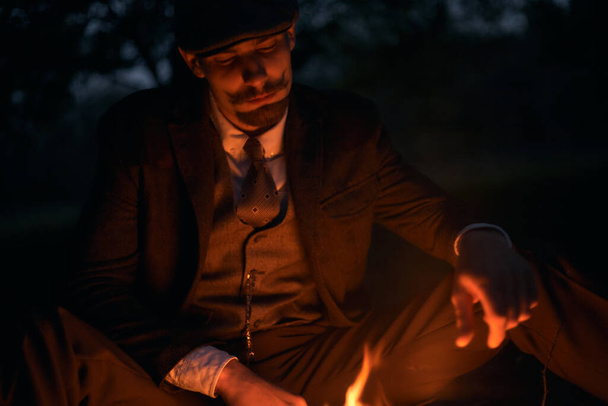 Porträt eines Mannes, der nachts in der Natur am Feuer sitzt. Filmreifes Oldtimer-Foto. Retro-Porträt eines Engländers mit Schiebermütze. - Foto, Bild