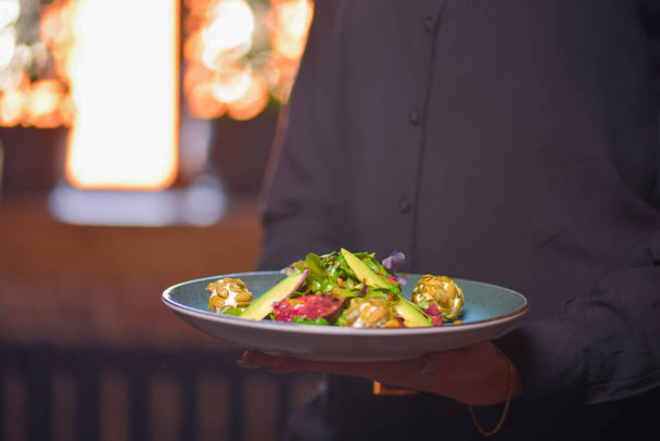 Официант в черной форме, рубашка, с заказанным здоровым шпинатом и салатом из рукколы с авокадо
. - Фото, изображение