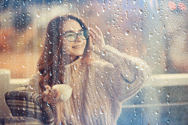 autumn coffee on a rainy day, girl behind a glass with a cup of hot coffee - Фото, зображення