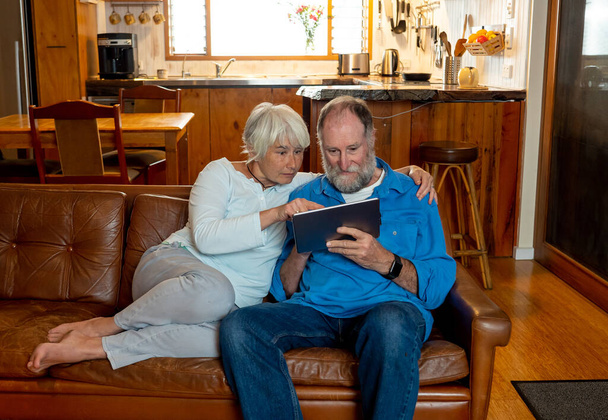 Happy active senior man and woman video calling family using digital tablet. Couple enjoying together watching stream movie or news on the internet on cozy vintage sofa. Coronavirus and life at home. - 写真・画像