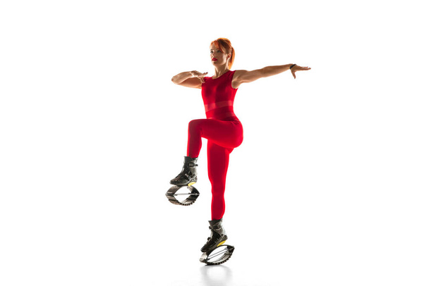 Beautiful redhead woman in a red sportswear jumping in a kangoo jumps shoes isolated on white studio background. - Foto, Imagem