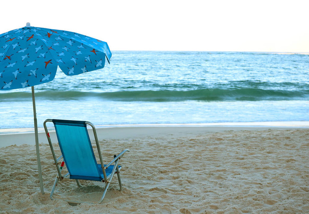 Пустое синее пляжное кресло и пляжный зонтик на песчаном пляже с видом на разбивающиеся волны в голубом океане
 - Фото, изображение