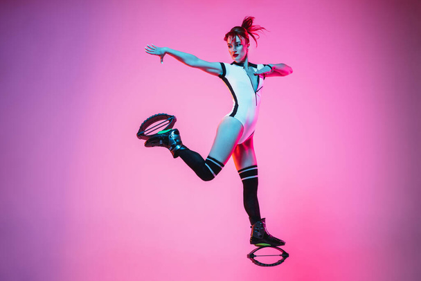 Beautiful redhead woman in a white sportswear jumping in a kangoo jumps shoes isolated on gradient studio background in neon light - Photo, image