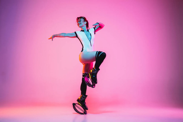 Beautiful redhead woman in a white sportswear jumping in a kangoo jumps shoes isolated on gradient studio background in neon light - Photo, Image