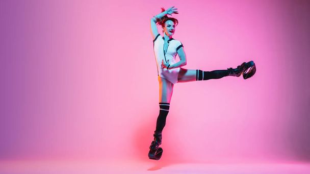 Beautiful redhead woman in a white sportswear jumping in a kangoo jumps shoes isolated on gradient studio background in neon light - Fotó, kép