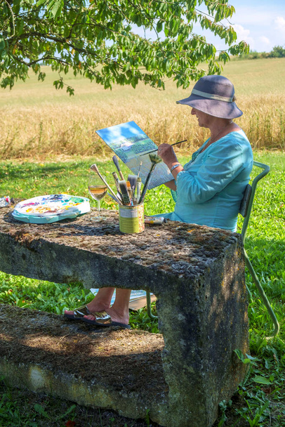 Eine Künstlerin sitzt am späten Nachmittag im Schatten mit Blick auf die ländliche Landschaft und arbeitet an dem Bild, mit dem sie früher am Tag begonnen hat. - Foto, Bild