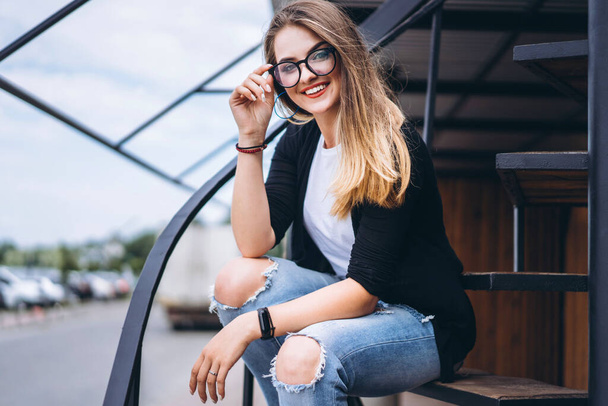 Beautiful girl with long hair and glasses sitting on metal stairs on the wooden background of house with vertical boards. Woman smiling and looking at camera. - Foto, afbeelding