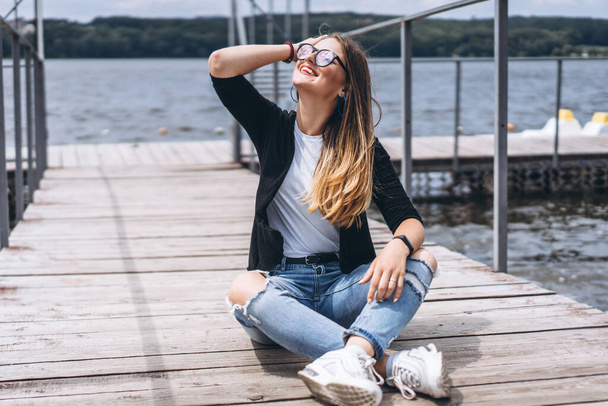 Young woman with long hair in stylish glasses posing on a wooden pier near the lake. Girl dressed in jeans and t-shirt smiling and looking away - Fotografie, Obrázek