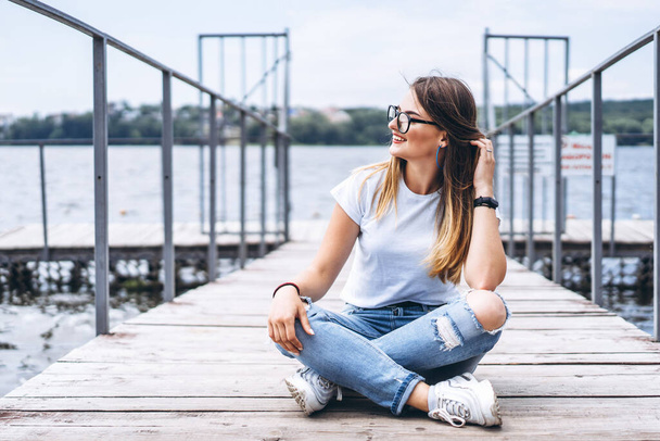 Young woman with long hair in stylish glasses posing on a wooden pier near the lake. Girl dressed in jeans and t-shirt smiling and looking away - Zdjęcie, obraz