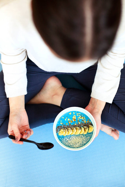 Высокий угол молодой женщины с миской здорового блюда улыбаясь и глядя на камеру, сидя скрестив ноги на полу после урока йоги. Закрыть
 - Фото, изображение