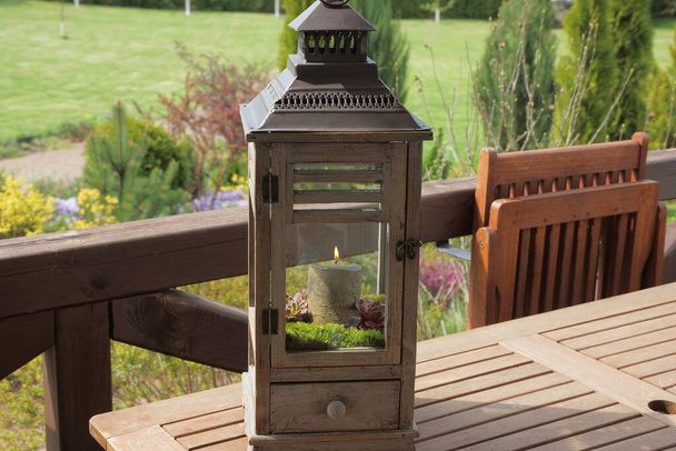 Szare drewniane pudełko ze szkłem i płonącą świecą wewnątrz stoi na brązowym stole na ulicy w ogrodzie z zielonym vegetatio - Zdjęcie, obraz