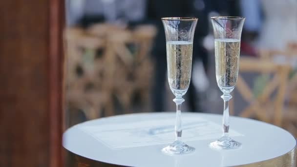 Een paar elegante champagneglazen staan op een rond bord van een kleine tafel voor gasten van een bruiloftsfeest en bankettafel, close shot. Bubbels lucht duiken op in volle glazen met gele mousserende wijn. - Video