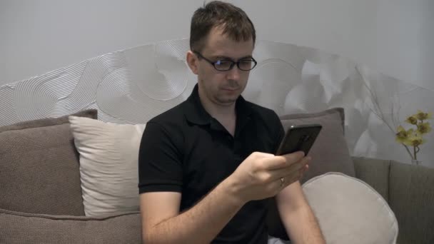 Seriózní muž v černém Polo tričku a brýlích sedí na gauči s mobilním telefonem - Záběry, video